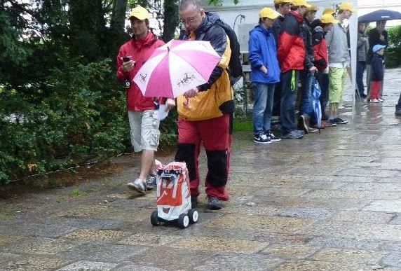 Robot v igelitové pláštěnce jede po mokré dlažbě, vedle něj kráčí člověk s růžovým deštníkem, v pozadí diváci v pláštěnkách.