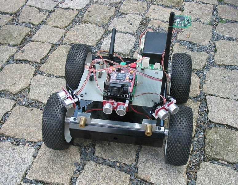 Robot ARBot ve verzi SRV1 na dlažbě: čtyřkolový podvozek, deska s elektronikou, kamera, tři sonary a dvě antény.