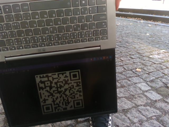 Snímek z pravé kamery robota: notebook otočený displejem dolů, na displeji QR kód, v pozadí dlažba.